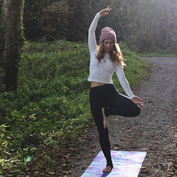 Woman practising yoga in a wooded area on a purple mat. The sun shines around her. 