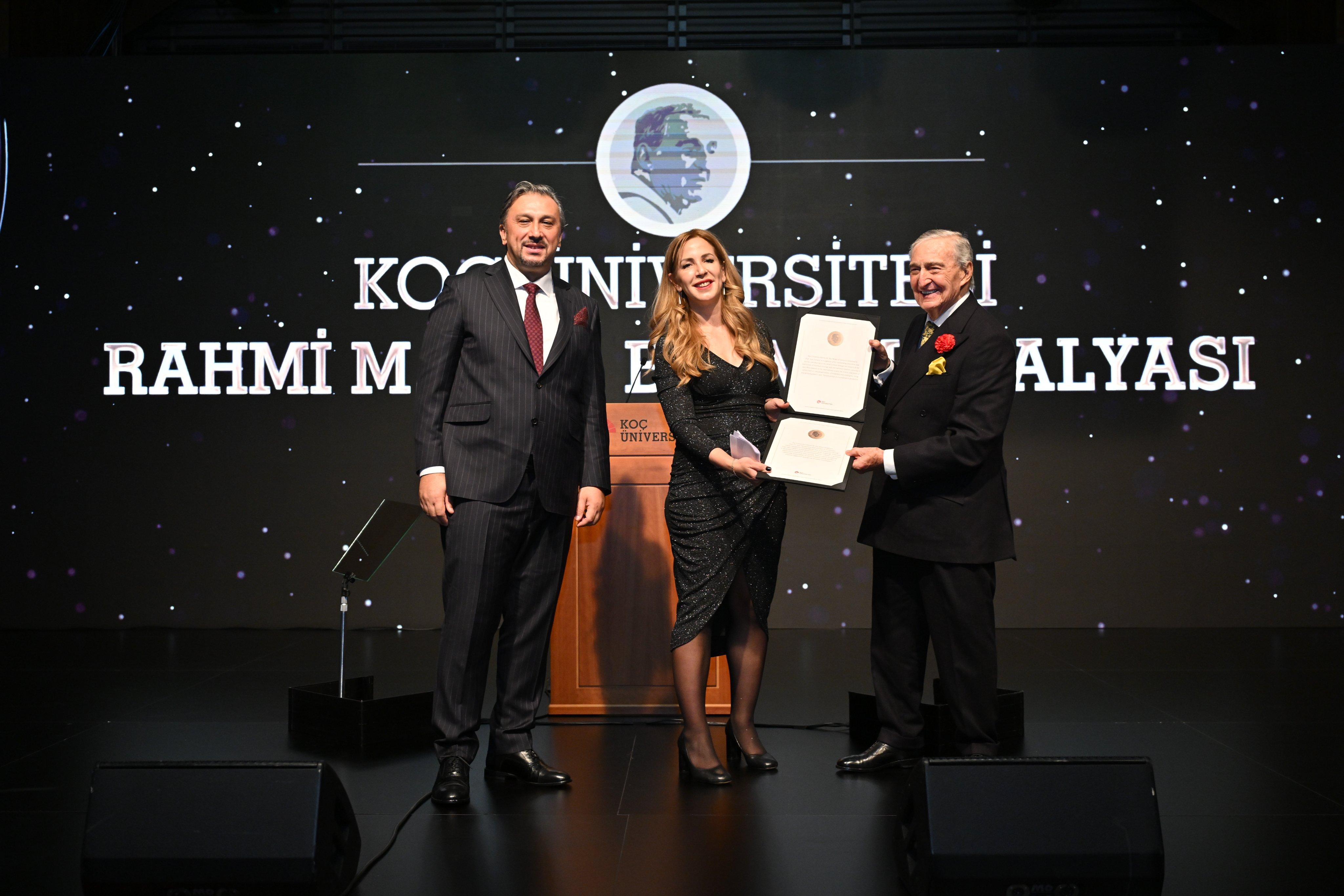 Professor Ayse Zarakol in black dress being awarded the Rahmi M. Koç Medal of Science certificate with two formally dressed presenters